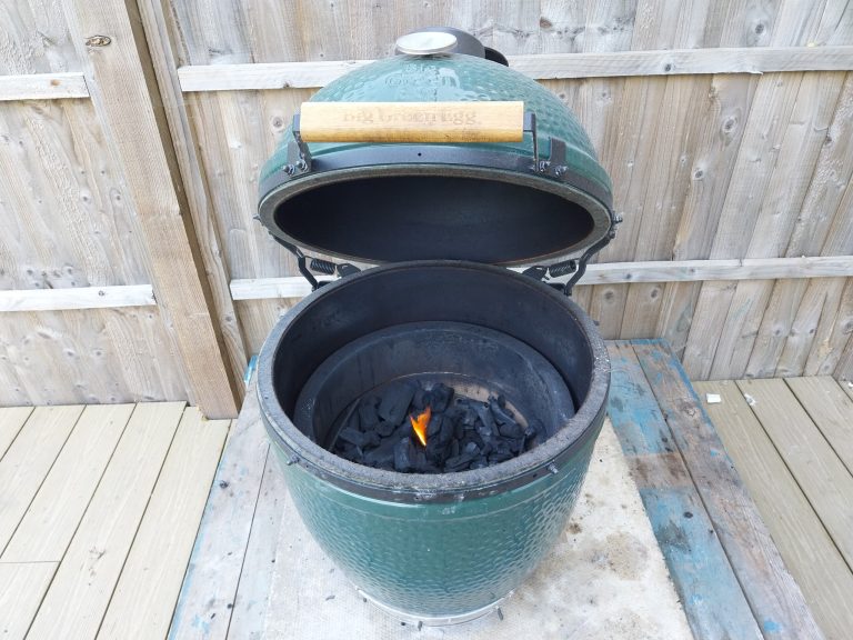 Big Green Egg Large with lid open, charcoal lit minion-style — single small flame visible at the front of the firebox, fence backdrop