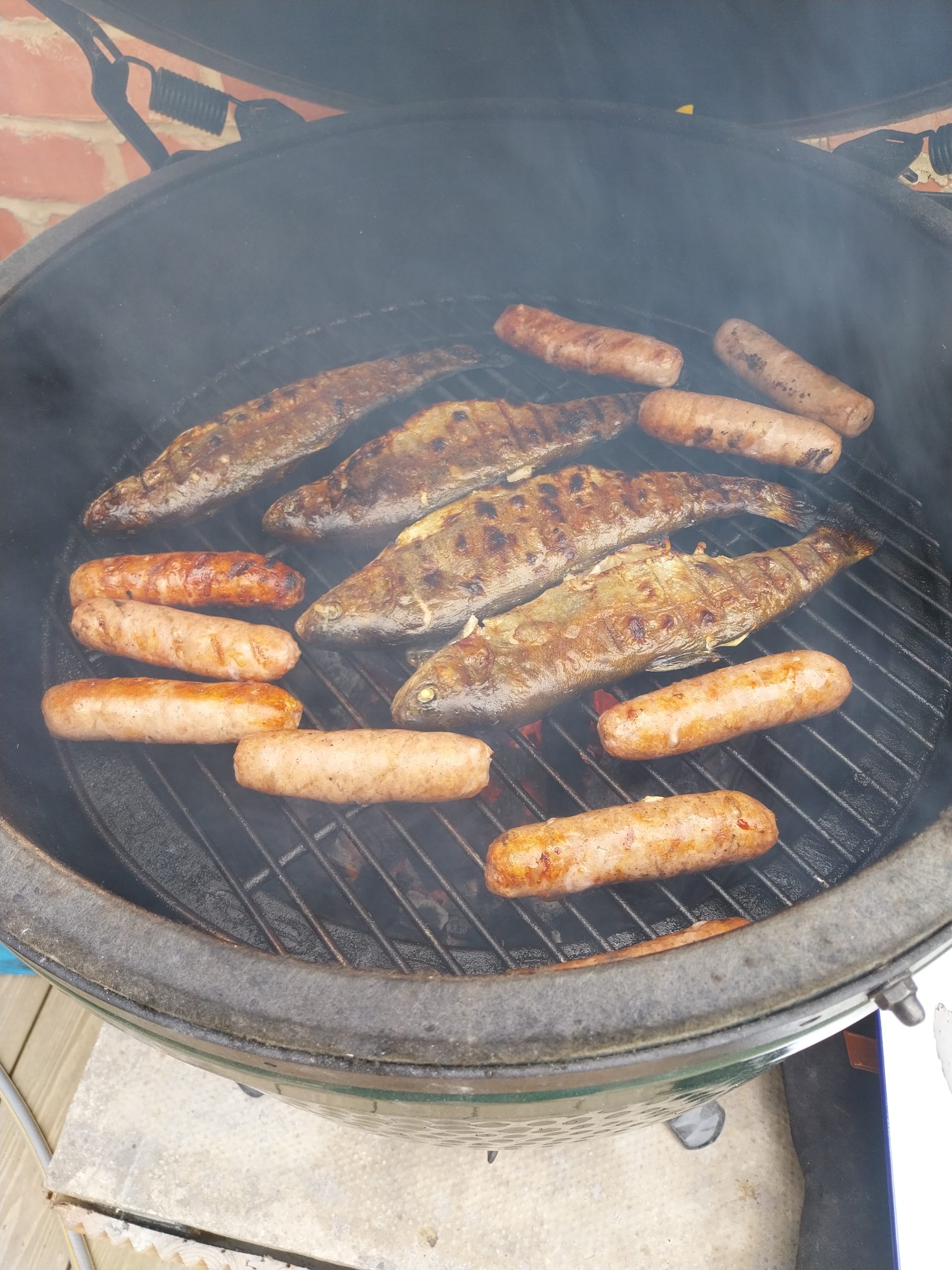 Whole rainbow trout cooking on the BGE Large grate alongside a small lamb rump, oiled skin glistening, dome held at 200°C direct — caught at Tillingbourne Farm on Friday 29 May 2026, cooked Sunday 31 May 2026