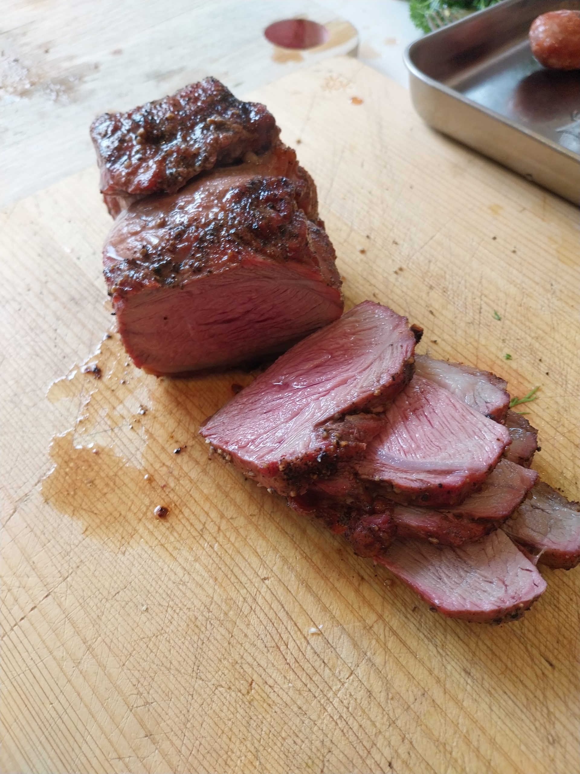 Sliced lamb rump on a wooden board, alternate angle — cherry-pink medium-rare interior with the rendered fat cap visible along the edges, second piece sitting behind