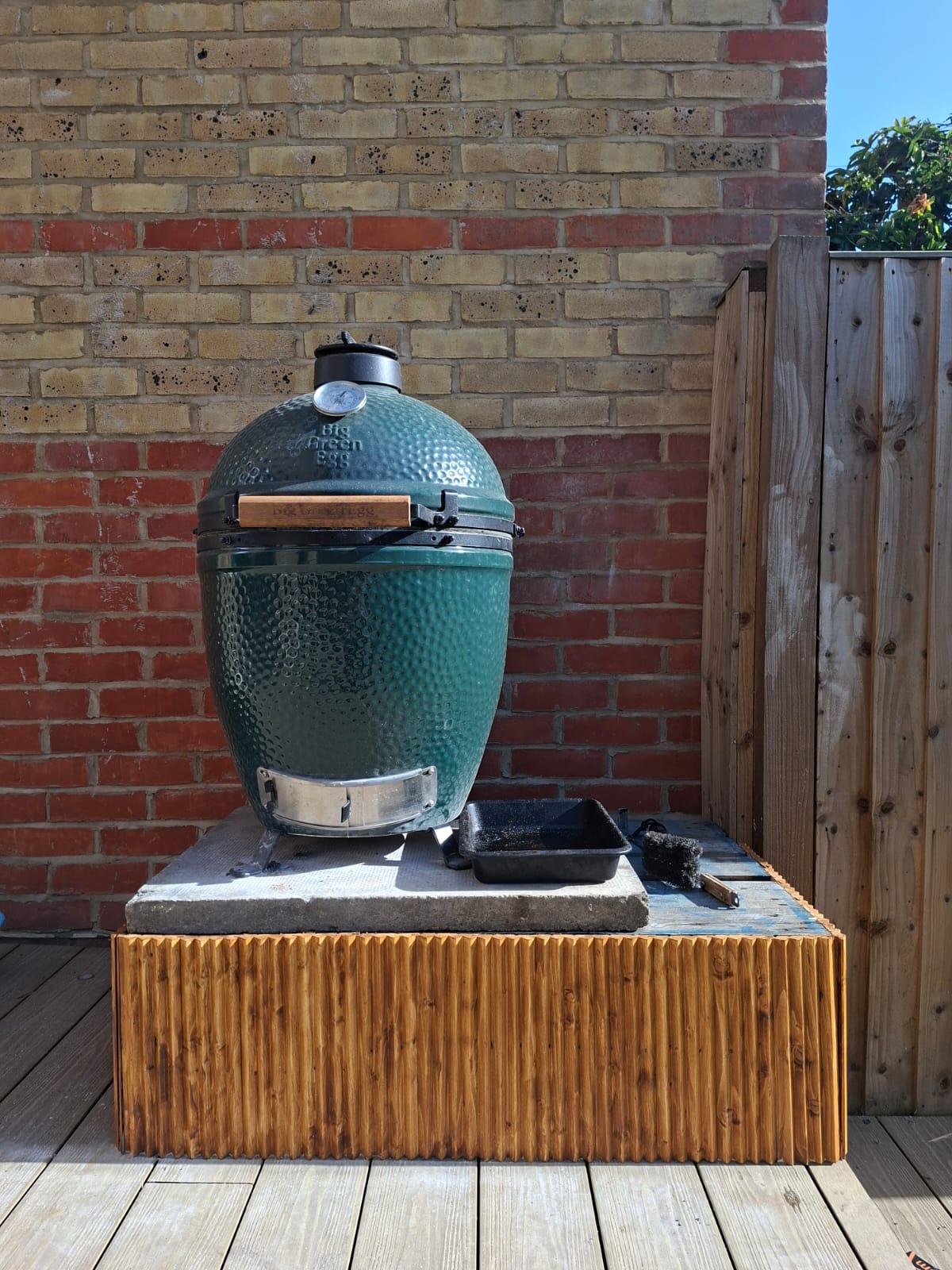 The Big Green Egg Large on its trimmed pallet base in a London garden — pallet cut in half, paving slab on top, painted wooden trim nail-gunned around the edges, photographed 22 May 2026.