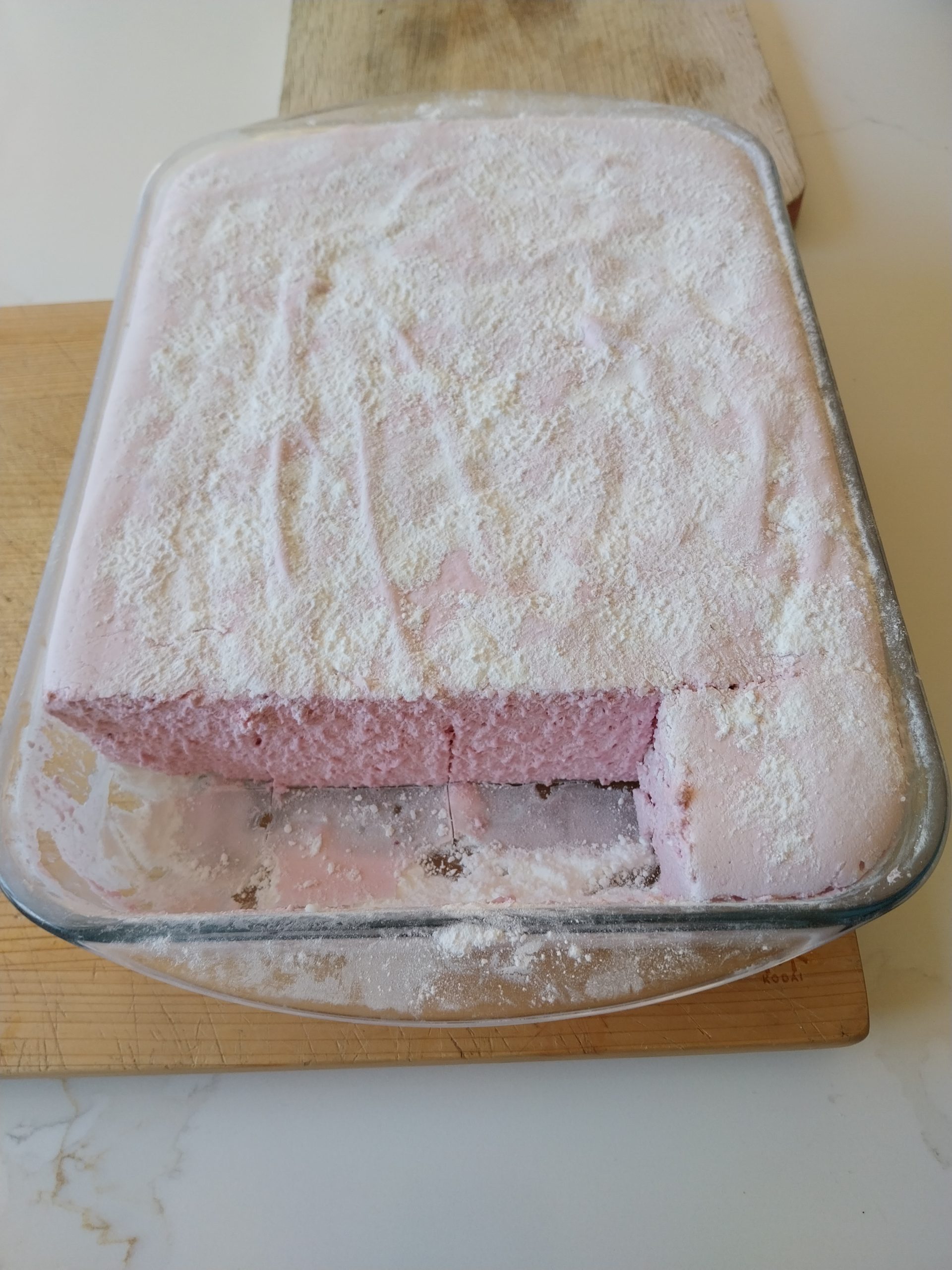 A slab of pale pink blackberry marshmallow set in a clear glass dish, heavily dusted with the icing sugar and cornflour mix, with one corner cut away showing an even aerated crumb right through