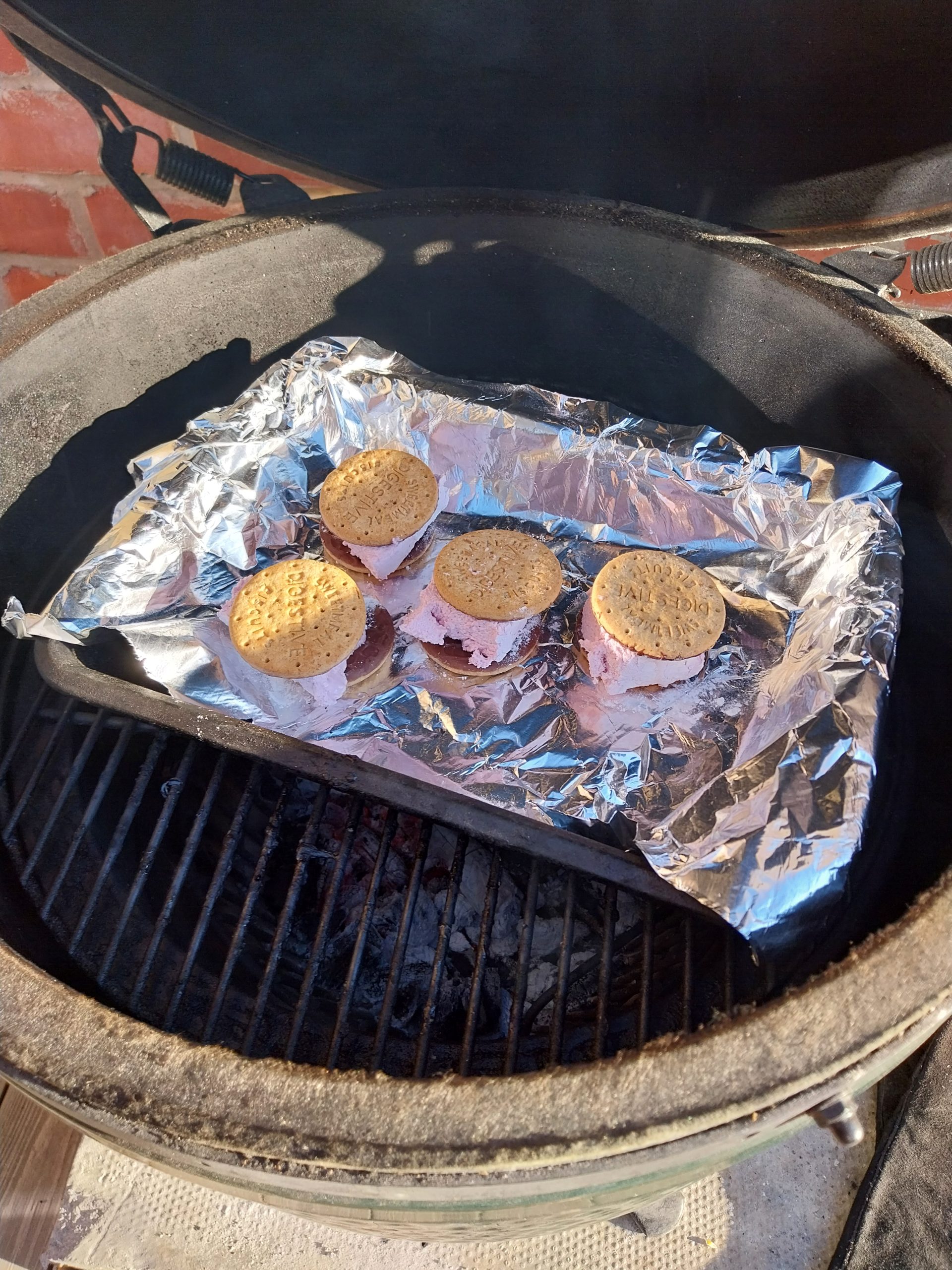 Four s'mores on foil on the cooking grid of a Big Green Egg Large, the blackberry marshmallow starting to slump out of the sides