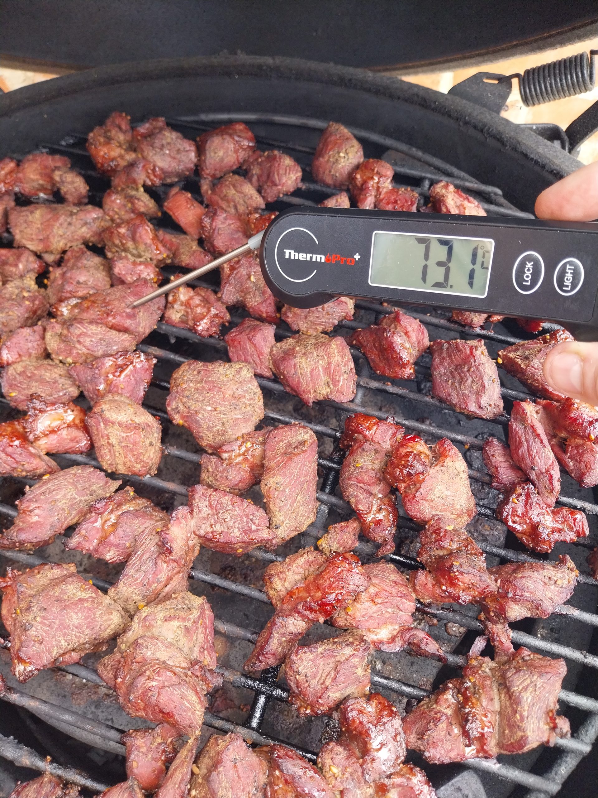 An instant-read thermometer reading 73.1°C in a cube of beef chuck on the kamado grate, bark set and dark after an hour and ten of smoke