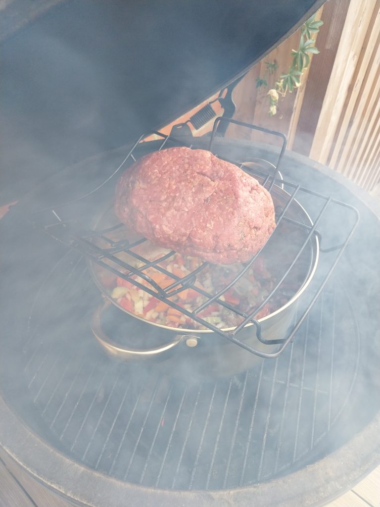 A seasoned beef mince ball on a wire rack standing over a stainless steel pan of softened peppers, onion, carrot and celery, on the cooking grate of a Big Green Egg Large in heavy smoke.