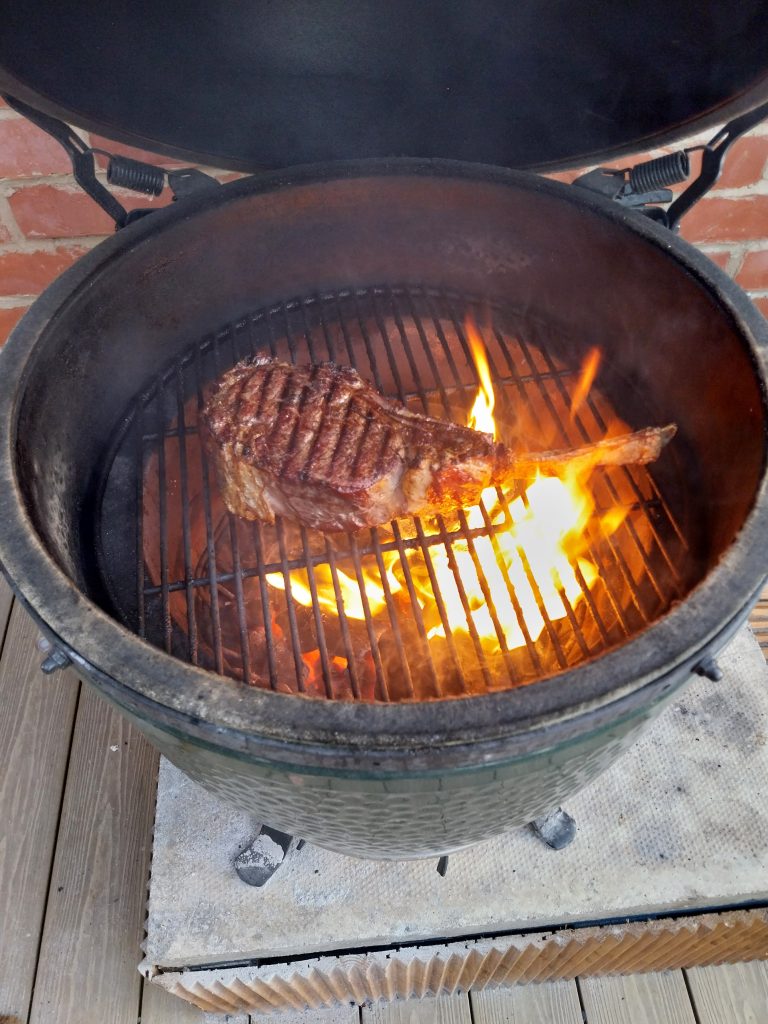 The tomahawk at the end of the sear on the Big Green Egg, lid open, with flames coming up through the grid under it.