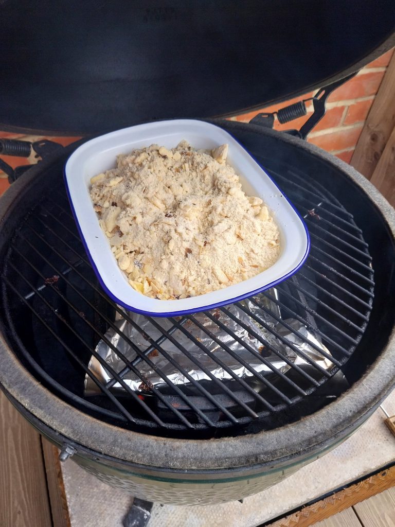 Apple crumble in a white blue-rimmed dish on the grid of my Big Green Egg Large, lid open, topping still pale before it browns