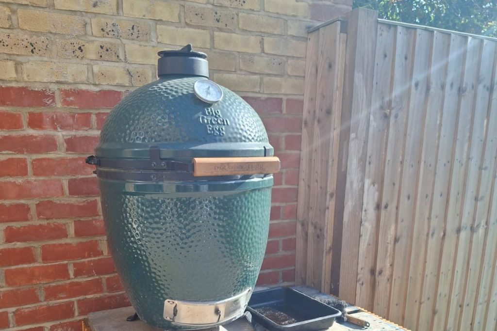 A Big Green Egg Large, lid closed, standing on a concrete slab against a brick wall, timber fence panels to one side.