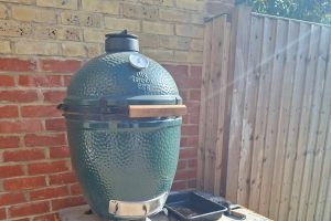 A Big Green Egg Large, lid closed, standing on a concrete slab against a brick wall, timber fence panels to one side.