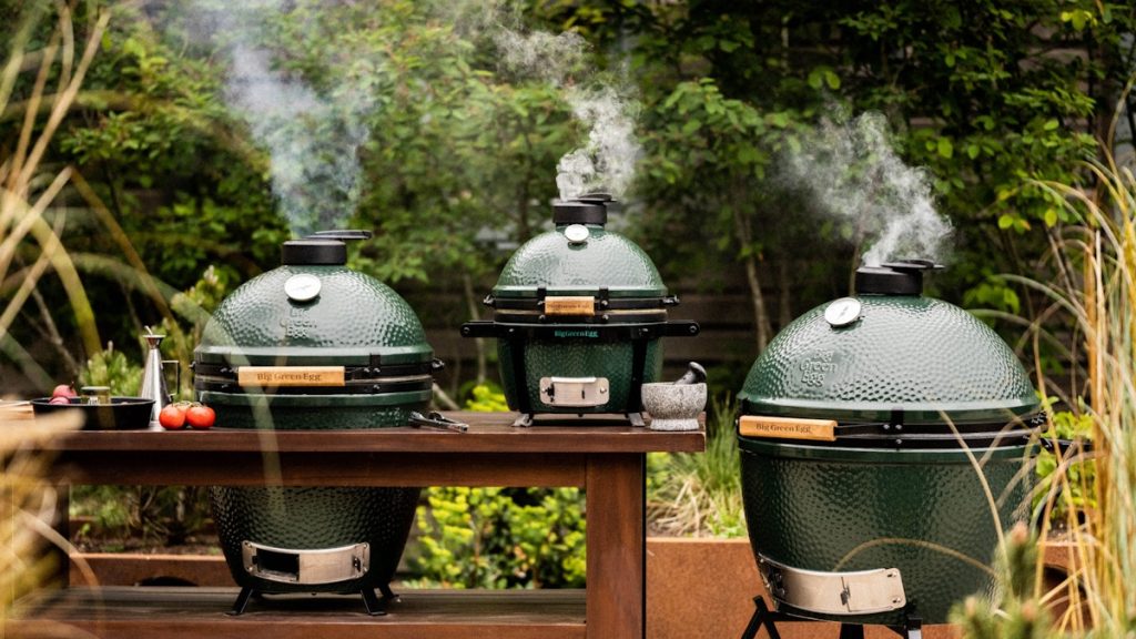 Three Big Green Eggs in a garden with smoke coming from their tops, two set into a wooden table and the biggest on a stand of its own.