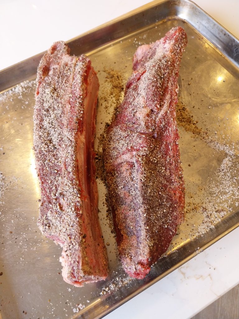 Two beef short ribs covered in a salt, pepper and garlic rub, on a metal tray before the cook