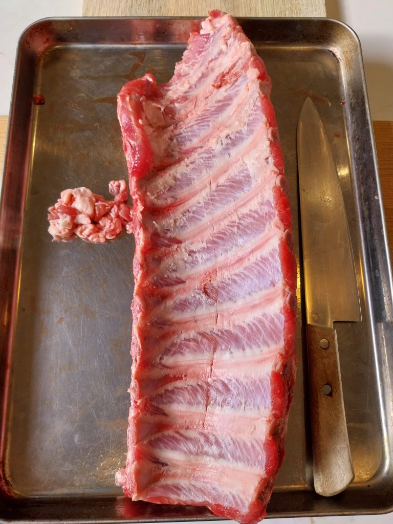 A raw rack of baby back ribs bone side up in a steel tray, the papery membrane pulled off and sitting beside it next to a knife