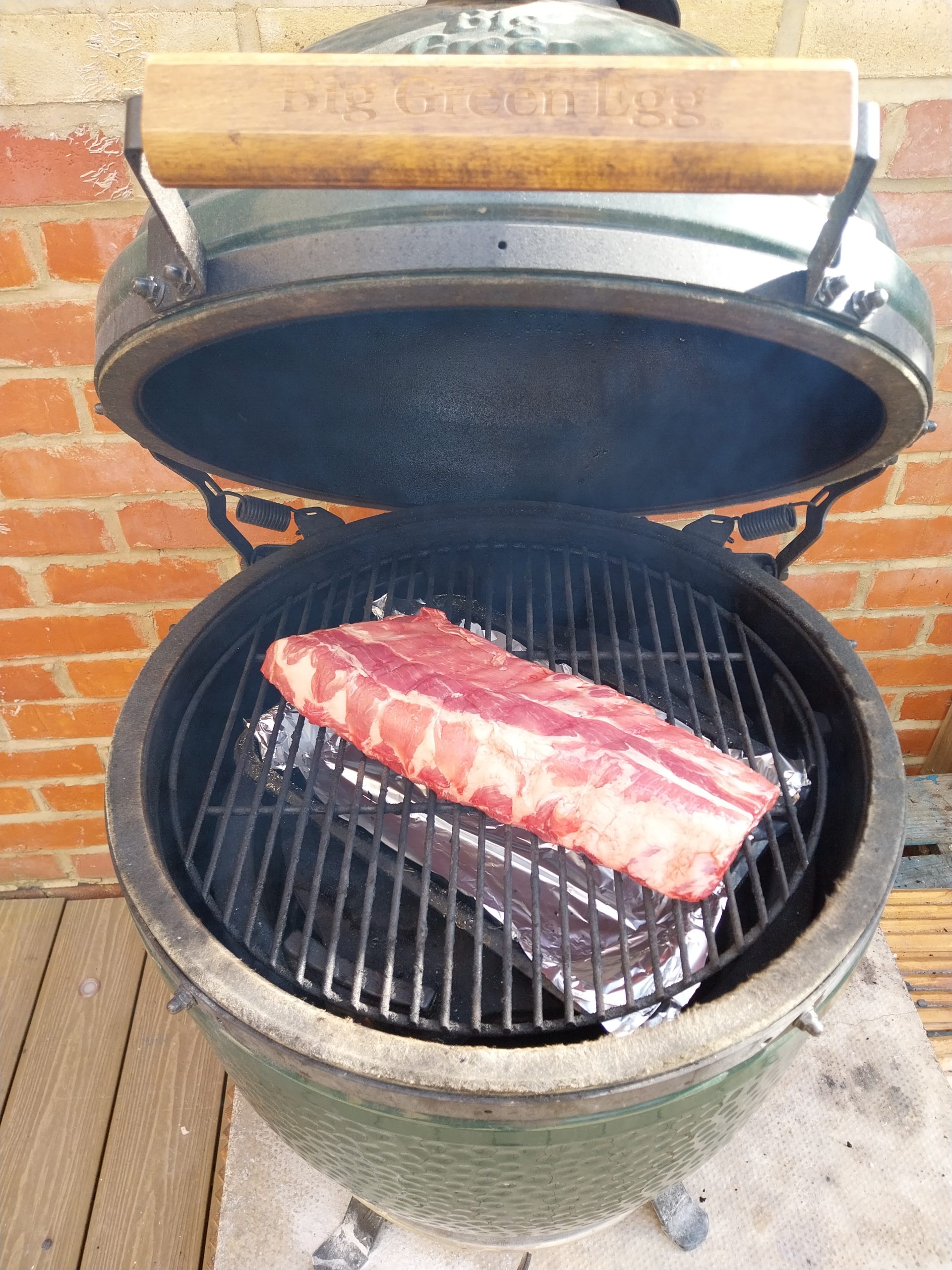 An unseasoned rack of baby back ribs going onto the grate of a Big Green Egg Large, with the foil-lined drip tray filling the space underneath