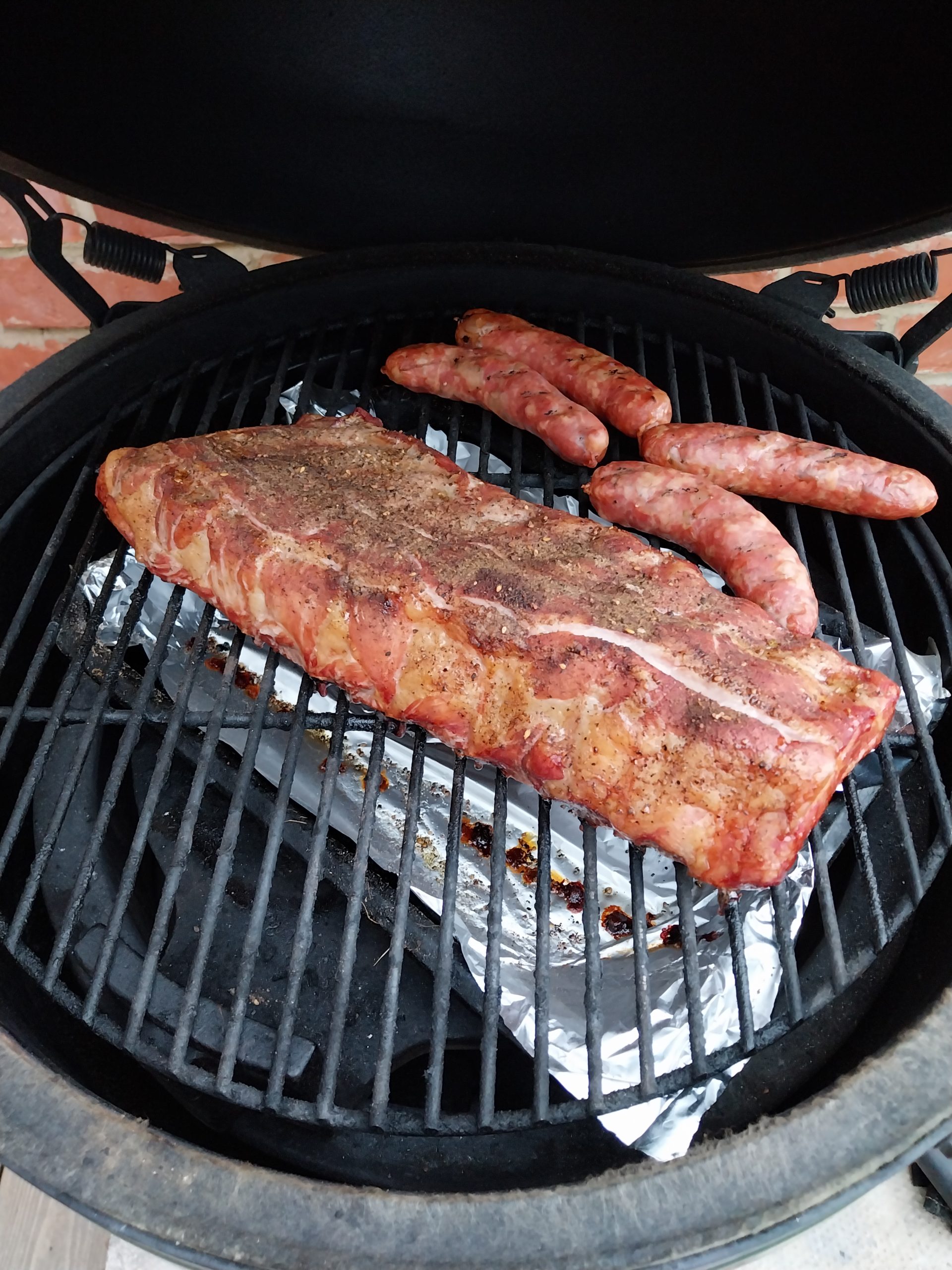 Two hours in: the ribs still pale with the bark not yet set, four fennel sausages on the grate beside them and the drip tray below