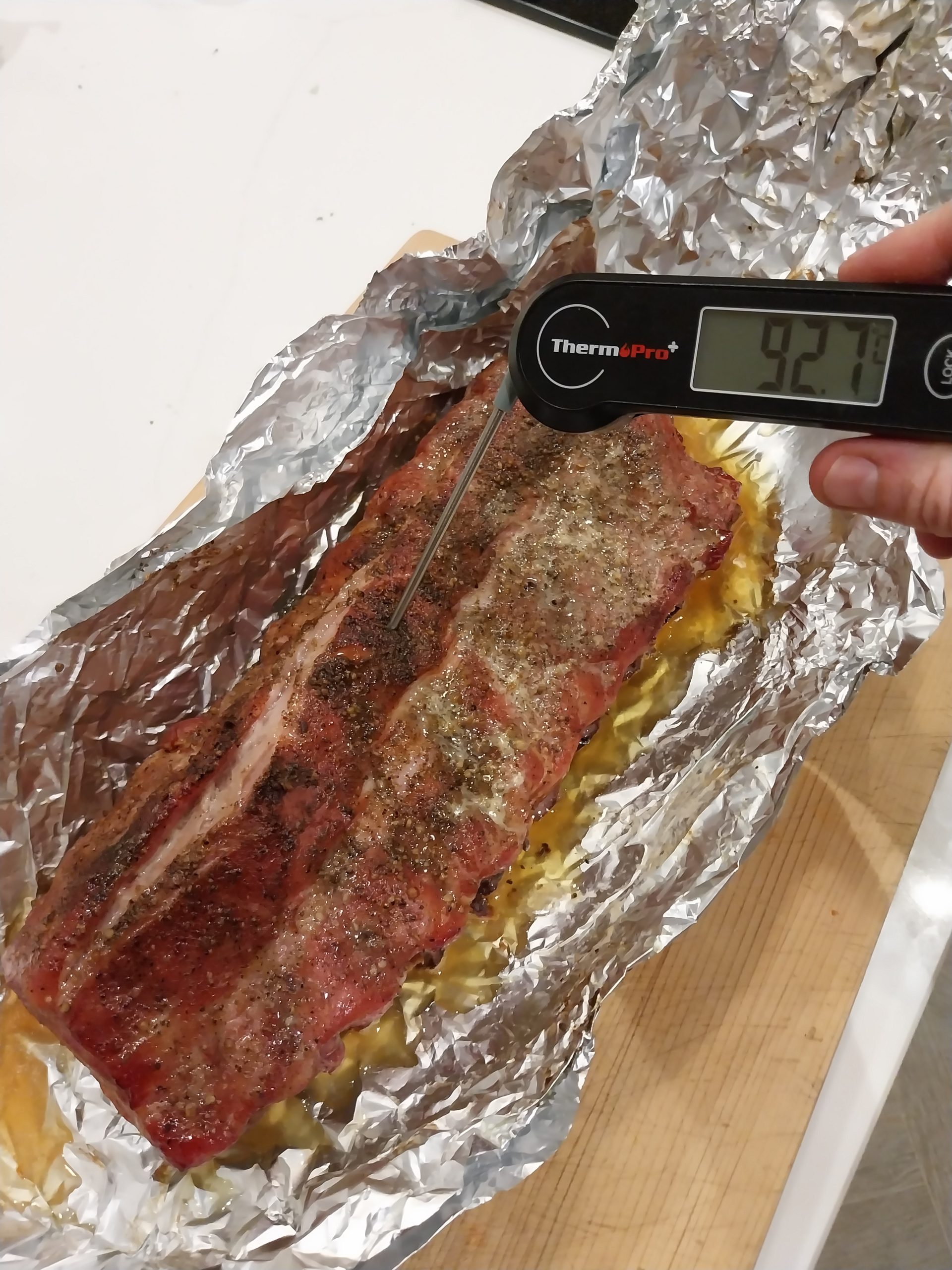 A ThermoPro instant-read pushed in between the bones showing 92.7°C, the unwrapped rack sitting in its opened foil on a board