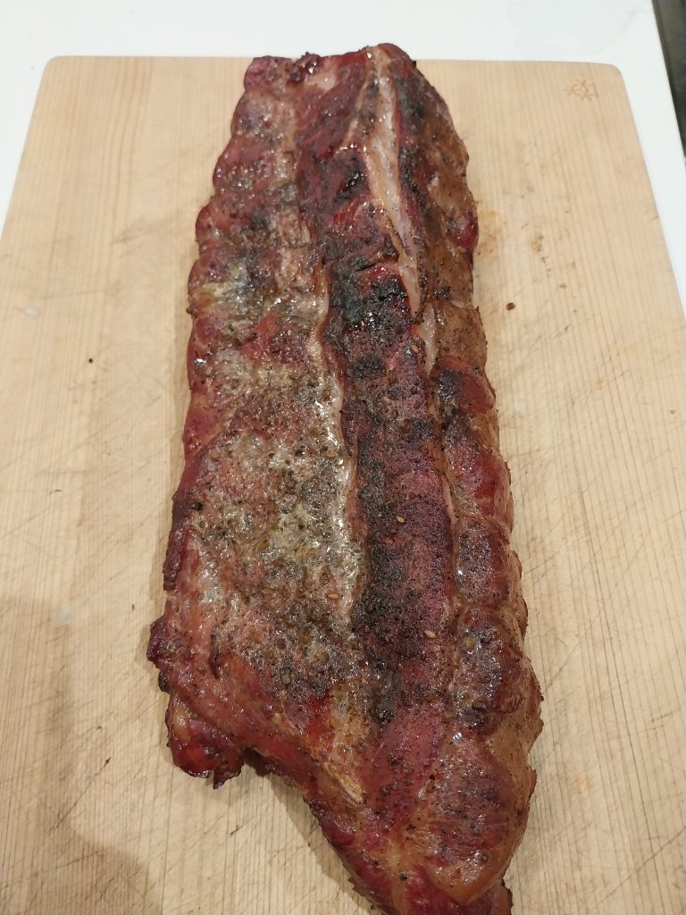 The finished rack of baby back ribs on a wooden board, meat side up, coarse black pepper still visible across a modest bark