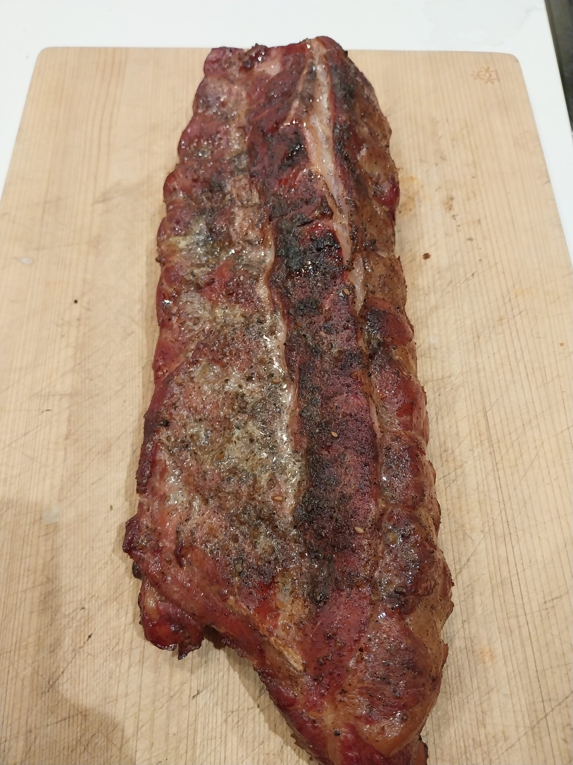 The finished rack of baby back ribs on a wooden board, meat side up, coarse black pepper still visible across a modest bark