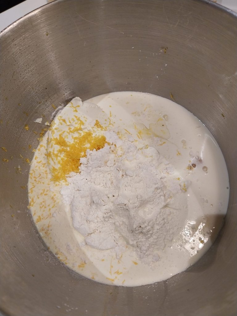 The cheesecake ingredients in a stand mixer bowl, with flour and lemon zest on top of the cream