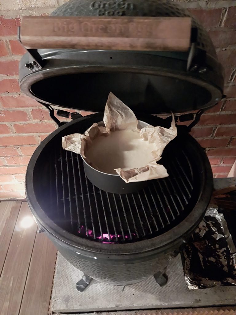 The cheesecake in a tin lined with baking parchment, sitting on the Big Green Egg grate with the lid open