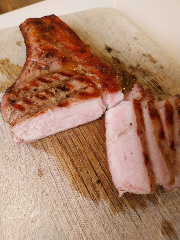 A thick bone-in pork chop on a wooden board, seared with grill marks, part carved and pink inside
