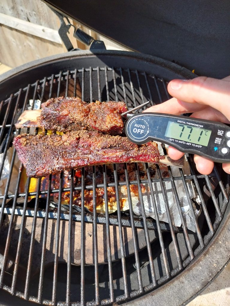Instant-read thermometer reading 77.1°C in the beef short rib with the fat cap left on, bark set, on the grate of the BGE Large at the wrap point of a 115°C indirect cook.
