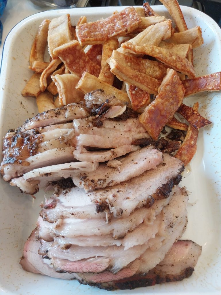 Slices of bone-in pork loin, pink in the middle, in a white enamel tray with a pile of crackling