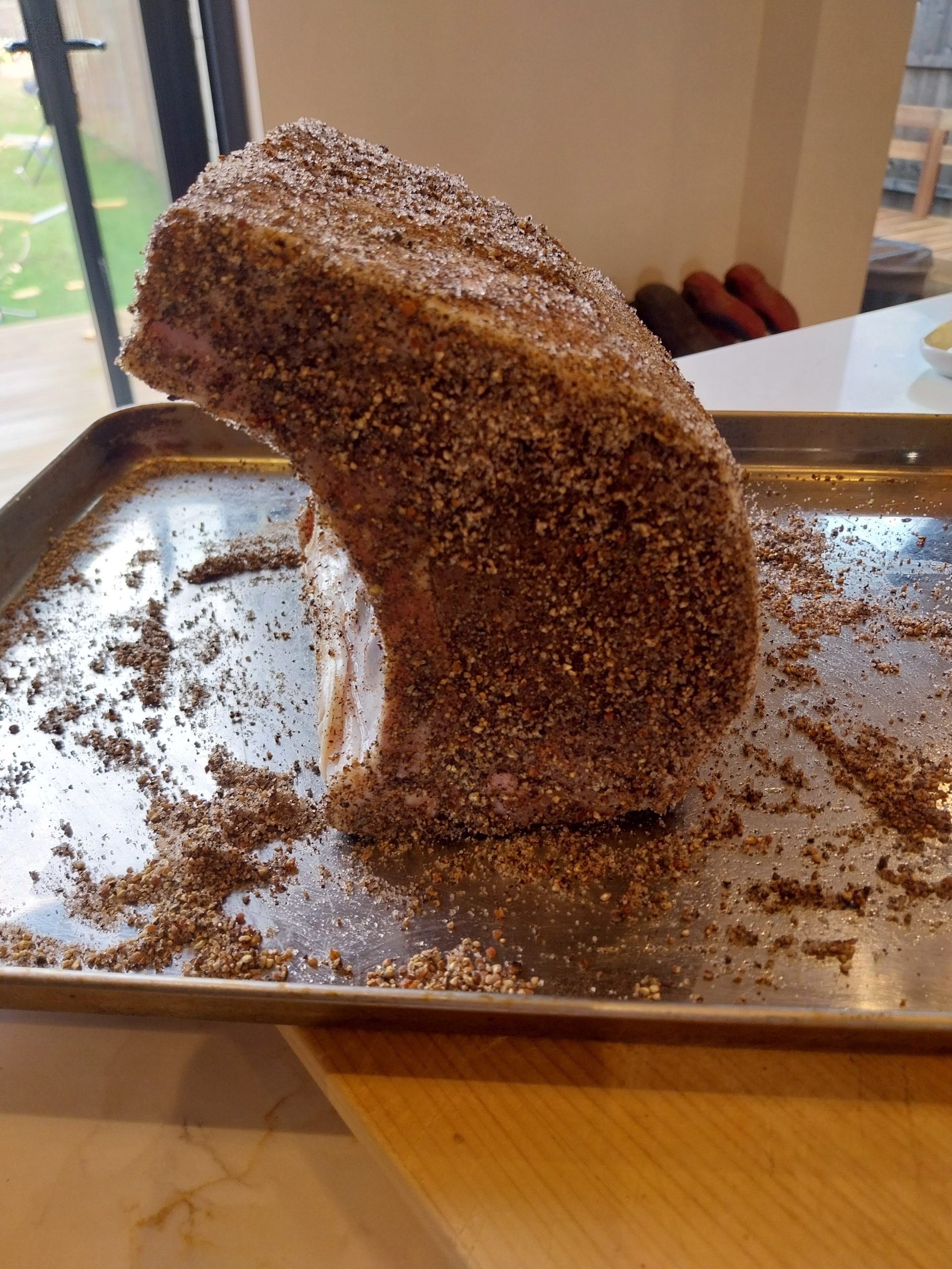 A bone-in pork loin coated in a coarse pepper rub, standing on a steel tray with loose rub around it