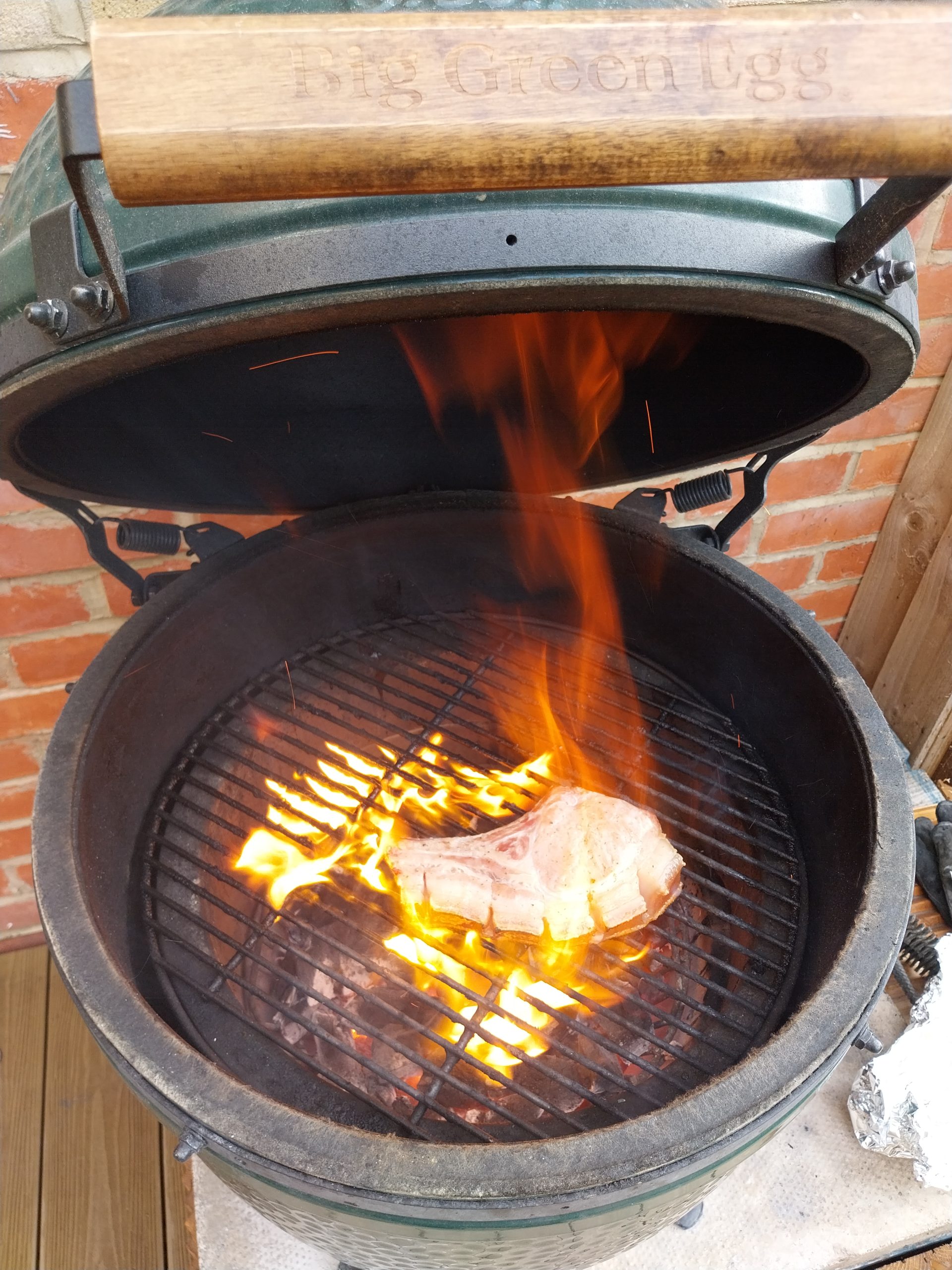 The pork chop searing over open coals on the Big Green Egg, with flames coming up from the rind
