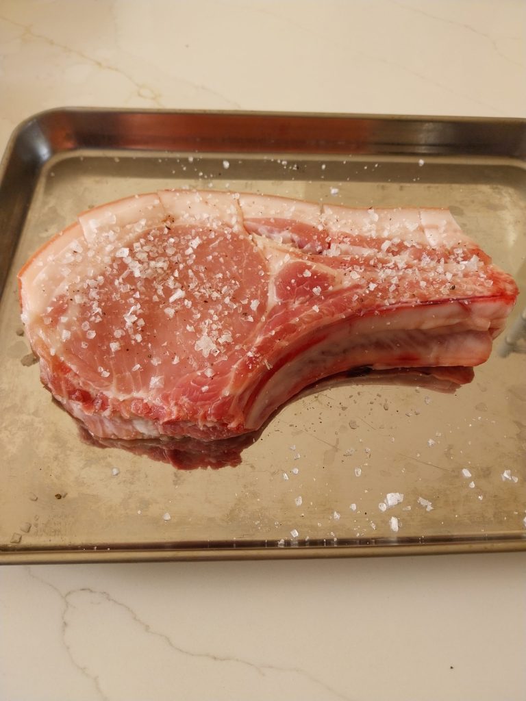 A raw bone-in Iberico pork chop on a metal tray, seasoned with salt, pepper and garlic