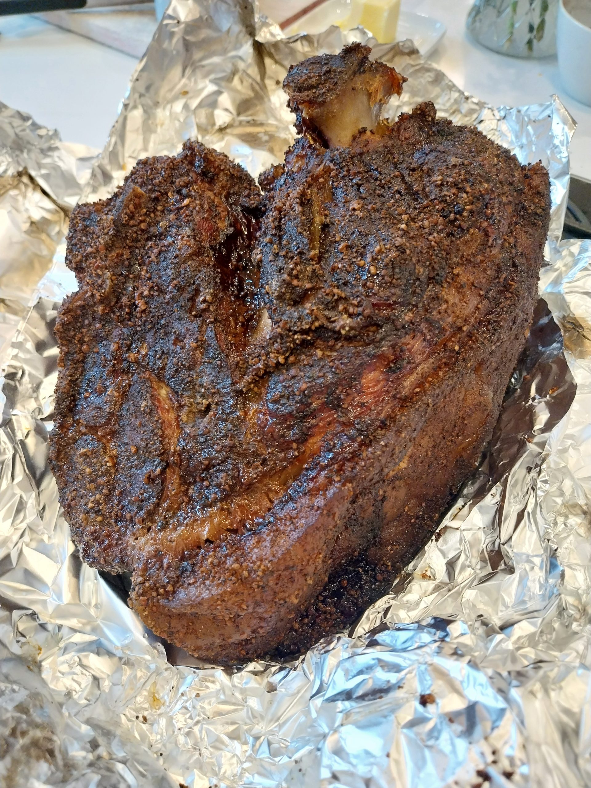 The whole cooked pork shoulder unwrapped on foil, with a dark peppery bark and the bone showing at the top