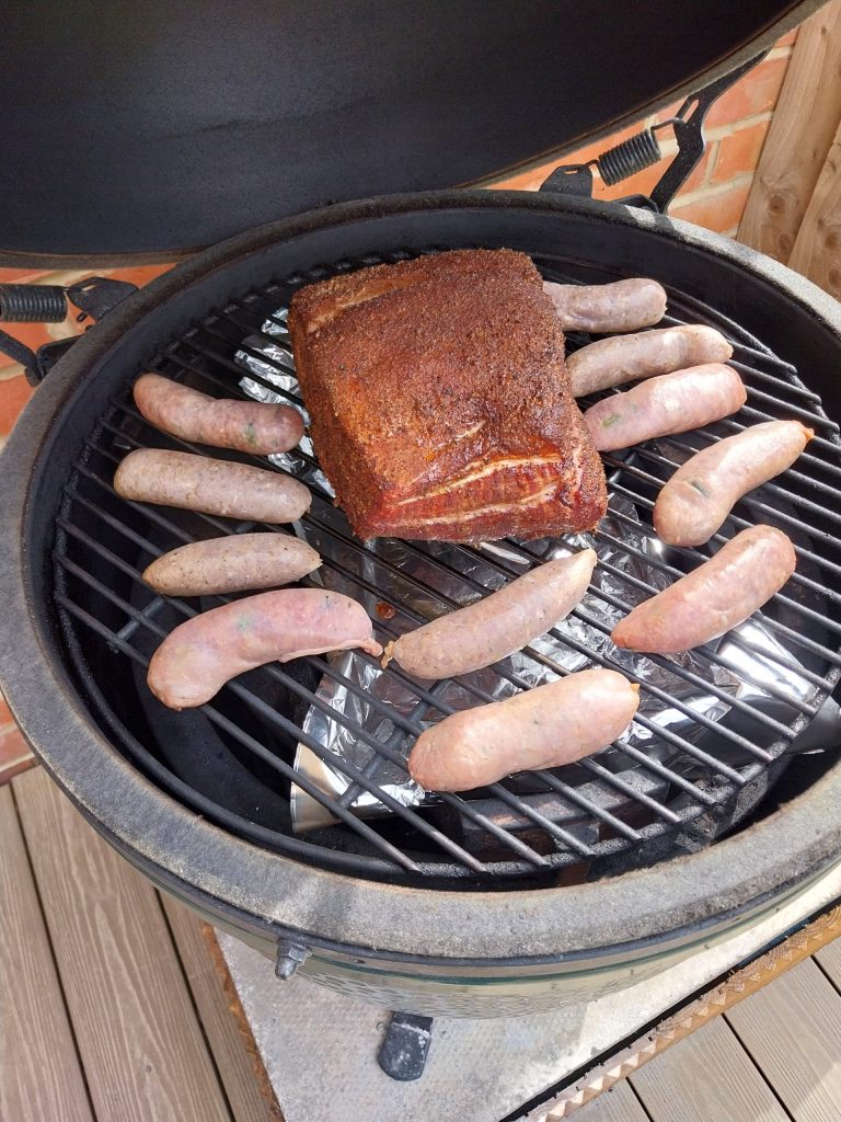 Raw sausages in a ring round a rubbed bone-in pork loin on the grid of a Big Green Egg, with a foil tray under the grate