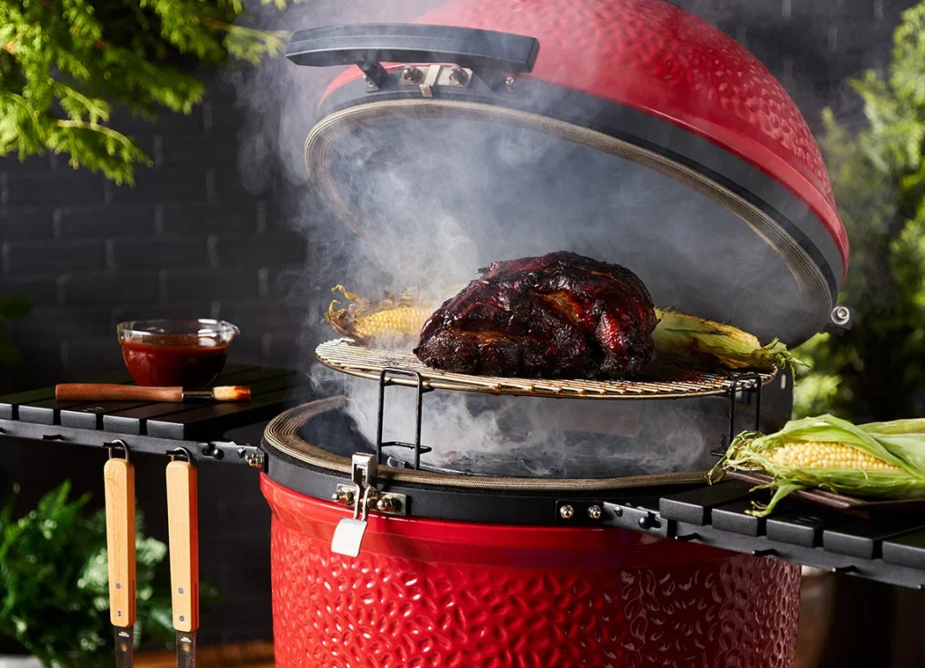 Kamado Joe Classic III — lid open and smoking, with a glazed joint on a raised rack and corn on the cob beside it. Image: Kamado Joe.