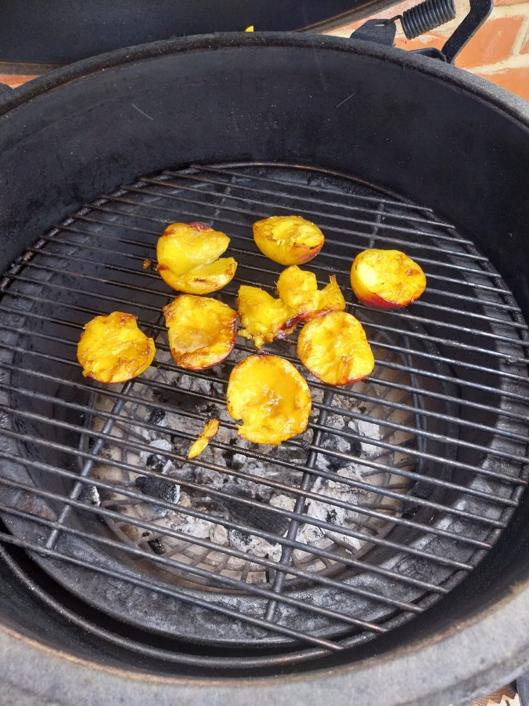 Eight peach halves cooking on the grid of the Big Green Egg over burnt-down ash, one half torn where the stone came out