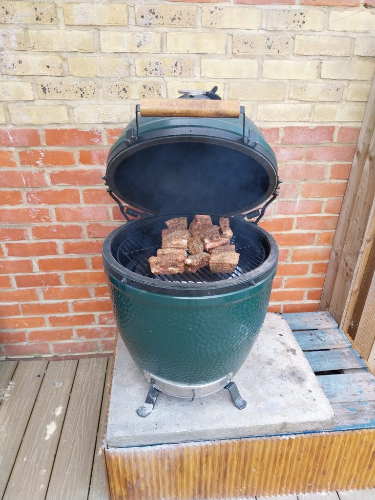Rubbed cubes of pork belly on the grid of a Big Green Egg Large, dome swung wide open with smoke still hanging inside it.