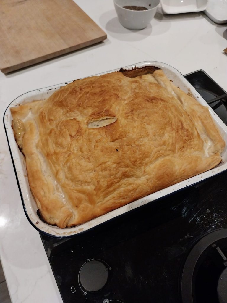 A chicken and bacon pie in a white enamel dish, the puff pastry lid risen and golden, with filling bubbling out at one edge
