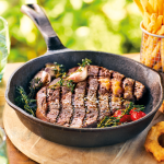 A griddled ribeye steak in a cast iron pan with herbs, garlic and tomatoes, from Donald Russell's ribeye listing.