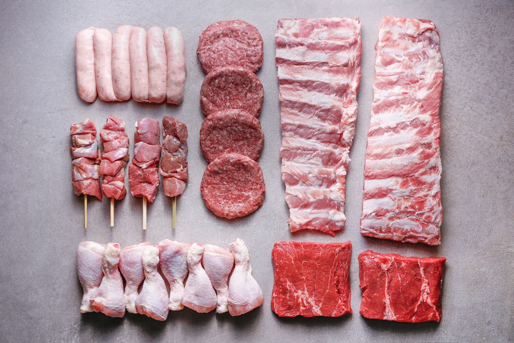 Raw BBQ meat laid out on grey slate: sausages, burgers, baby back ribs, lamb brochettes on skewers, chicken drumsticks and flat iron steaks, from HG Walter's Classic BBQ Box listing.