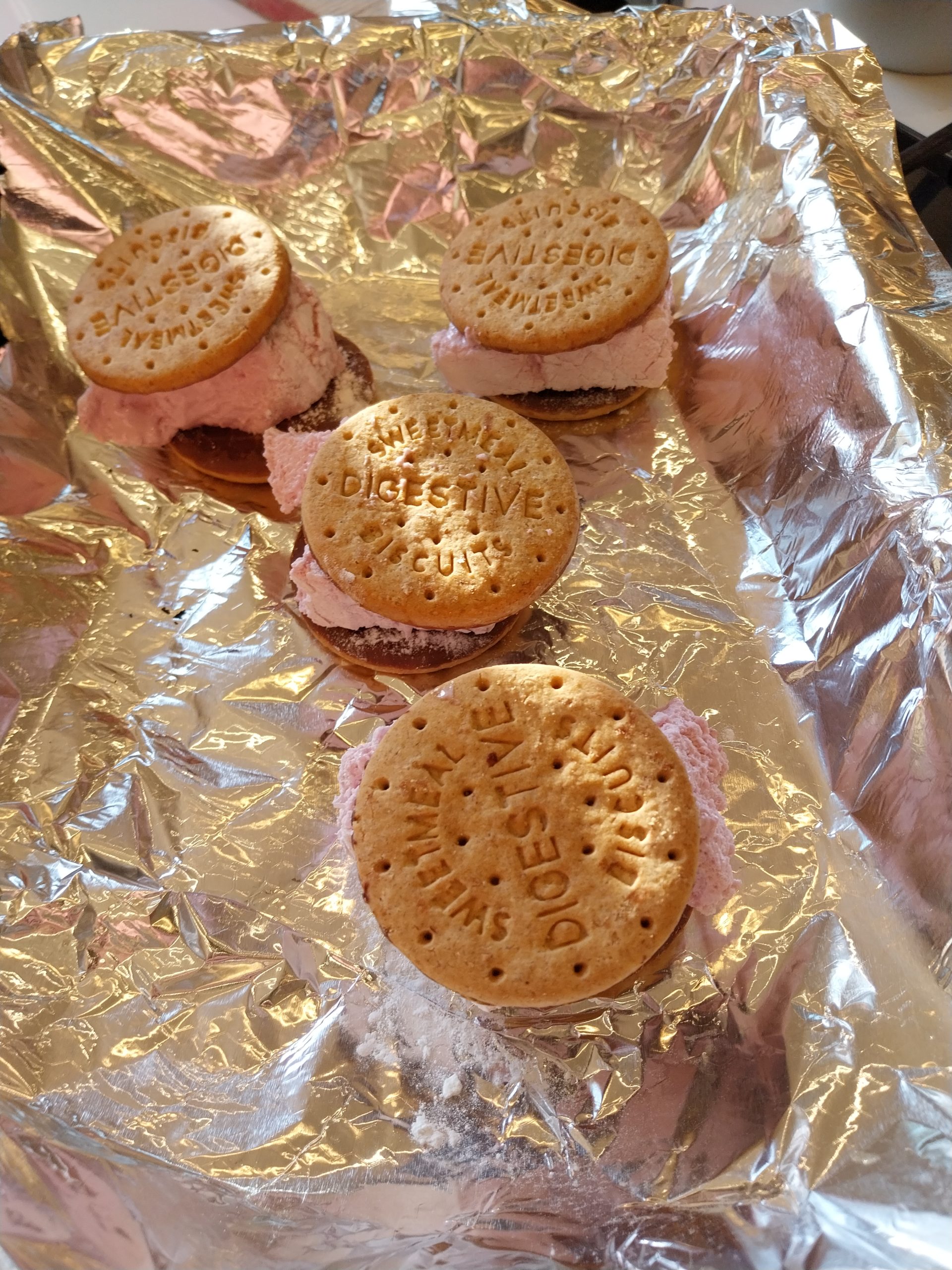 Four s'mores built on foil, a piece of pink blackberry marshmallow between two digestive biscuits