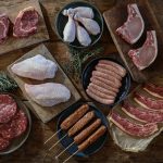 Raw meat laid out on a wooden table: ribeye steaks, chicken breasts and drumsticks, pork chops, sausages, burgers, skewers and thin-cut beef short ribs, from Swaledale's BBQ Meat Box listing.