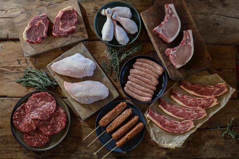 Raw meat laid out on a wooden table: ribeye steaks, chicken breasts and drumsticks, pork chops, sausages, burgers, skewers and thin-cut beef short ribs, from Swaledale's BBQ Meat Box listing.
