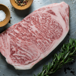 A raw A5 Japanese Wagyu sirloin steak, heavily marbled, on a dark board with rosemary, salt and peppercorns, from Tom Hixson's listing.
