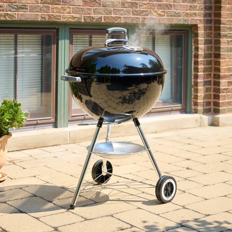 A black Weber Original Kettle 57cm on a brick patio with smoke rising from the lid.